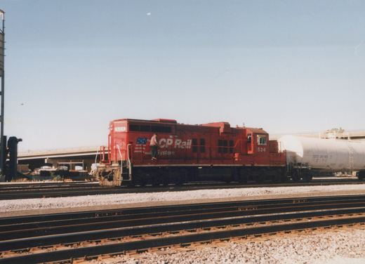 A red CP Rail locomotive number 534, with a person standing on its side, is parked in a multi-track rail yard next to a white tank car, with an overpass visible in the background.