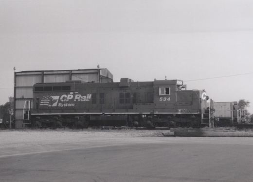 A black and white photograph of a CP Rail System diesel locomotive, number 534, standing in a railyard next to a building under an overcast sky.