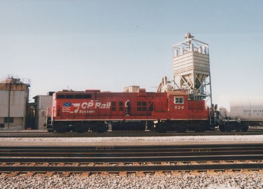A red diesel locomotive with 'CP Rail System' lettering and the number 534 is parked on tracks in a train yard, with one person standing on it and a white tank car nearby.