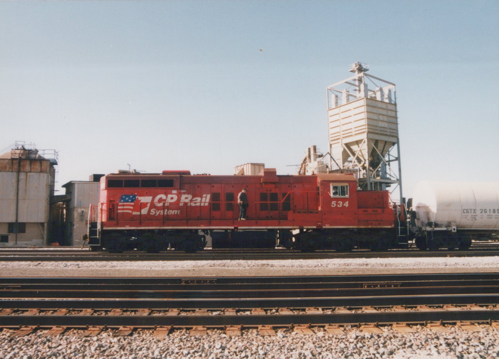 A red diesel locomotive with 'CP Rail System' lettering and the number 534 is parked on tracks in a train yard, with one person standing on it and a white tank car nearby.