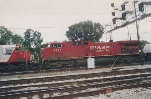 A red CP Rail System locomotive numbered 9554 is pictured on a railroad track, with a white and red work vehicle marked 6038 nearby, in a rail yard with multiple tracks and a signal bridge in the background.