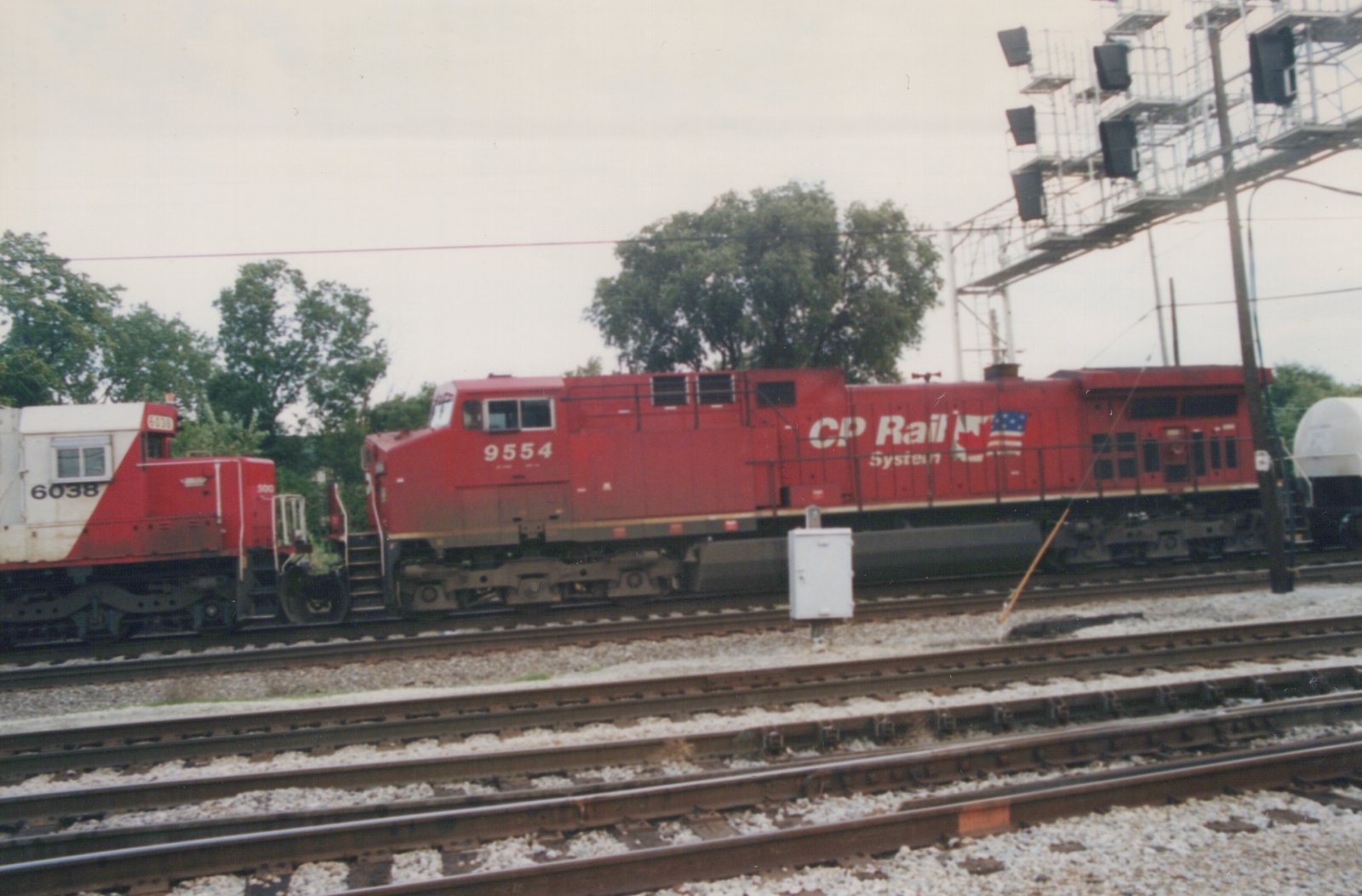 A red CP Rail System locomotive numbered 9554 is pictured on a railroad track, with a white and red work vehicle marked 6038 nearby, in a rail yard with multiple tracks and a signal bridge in the background.