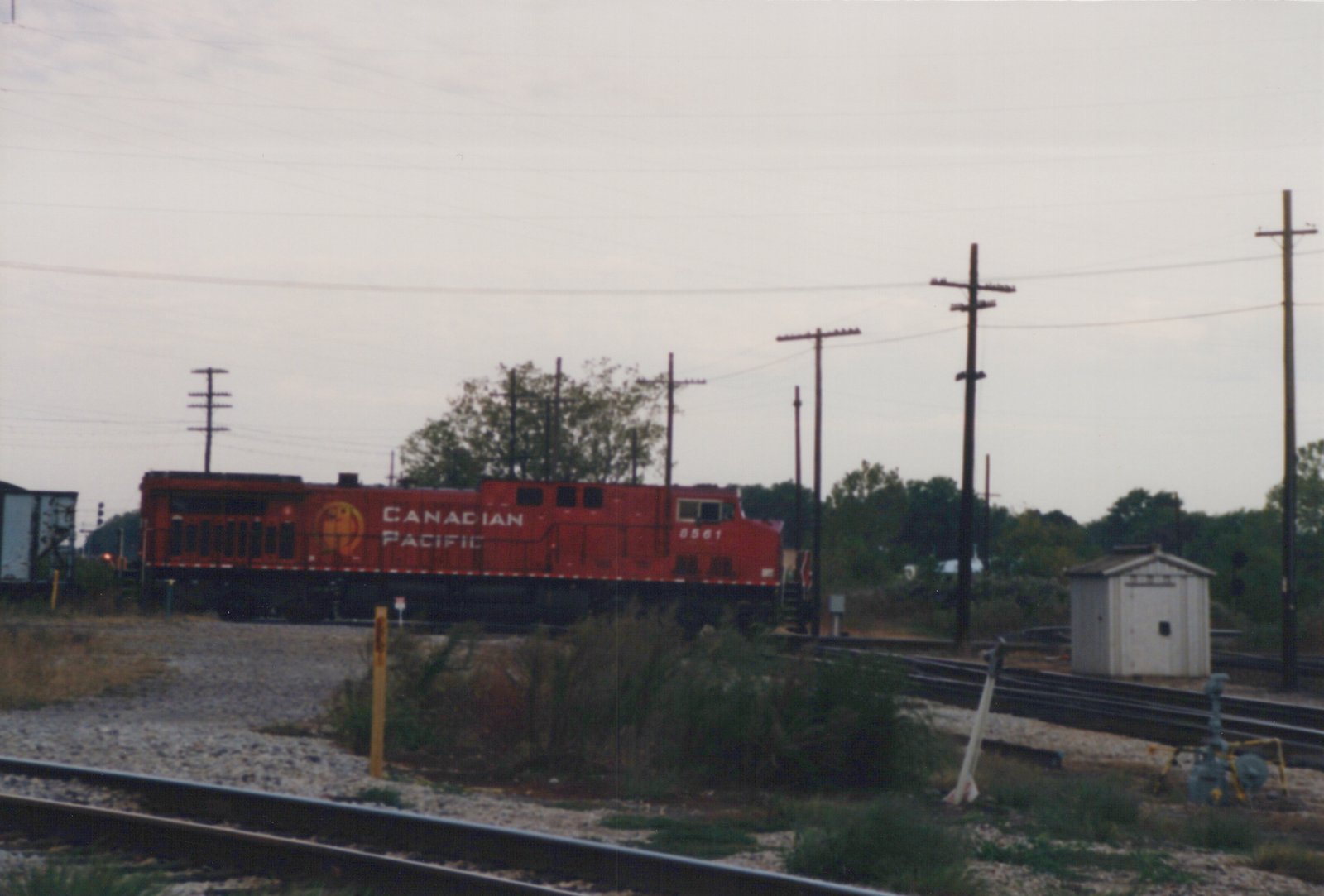 A red Canadian Pacific locomotive numbered 8561 sits on tracks in a yard with utility poles and a small shed nearby, under a gray sky.