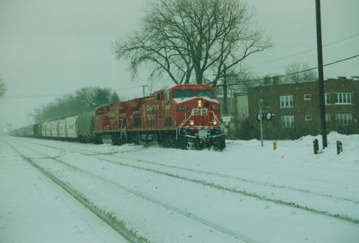 A red Canadian Pacific locomotive pulls a long line of freight cars on snow-covered rails beside a brick building and bare trees.