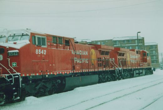 Two red Canadian Pacific diesel locomotives, numbers 8542 and 43, on snowy railroad tracks with buildings in the background.