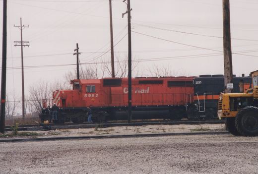 Side view of a red CP Rail locomotive numbered 5962 coupled to a black Indiana Rail Road locomotive; two people stand near the cab; bare trees and utility poles in background under an overcast sky.