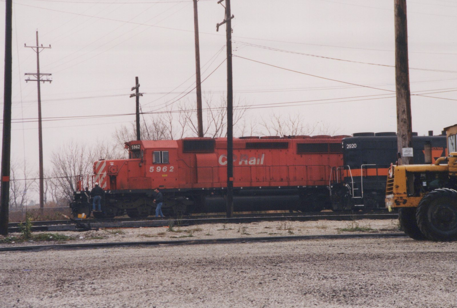 Side view of a red CP Rail locomotive numbered 5962 coupled to a black Indiana Rail Road locomotive; two people stand near the cab; bare trees and utility poles in background under an overcast sky.