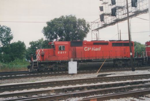 A red CP Rail diesel locomotive, number 5811, stands on a set of tracks in a rail yard with green trees in the background.