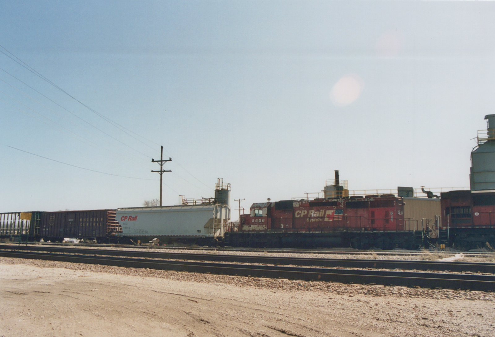 A red CP Rail System locomotive numbered 5606 stands in a railyard beside a white CP Rail freight car and a dark Wisconsin Central boxcar under a clear blue sky.