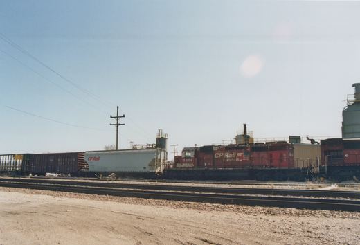 A red CP Rail System locomotive numbered 5606 stands in a railyard beside a white CP Rail freight car and a dark Wisconsin Central boxcar under a clear blue sky.