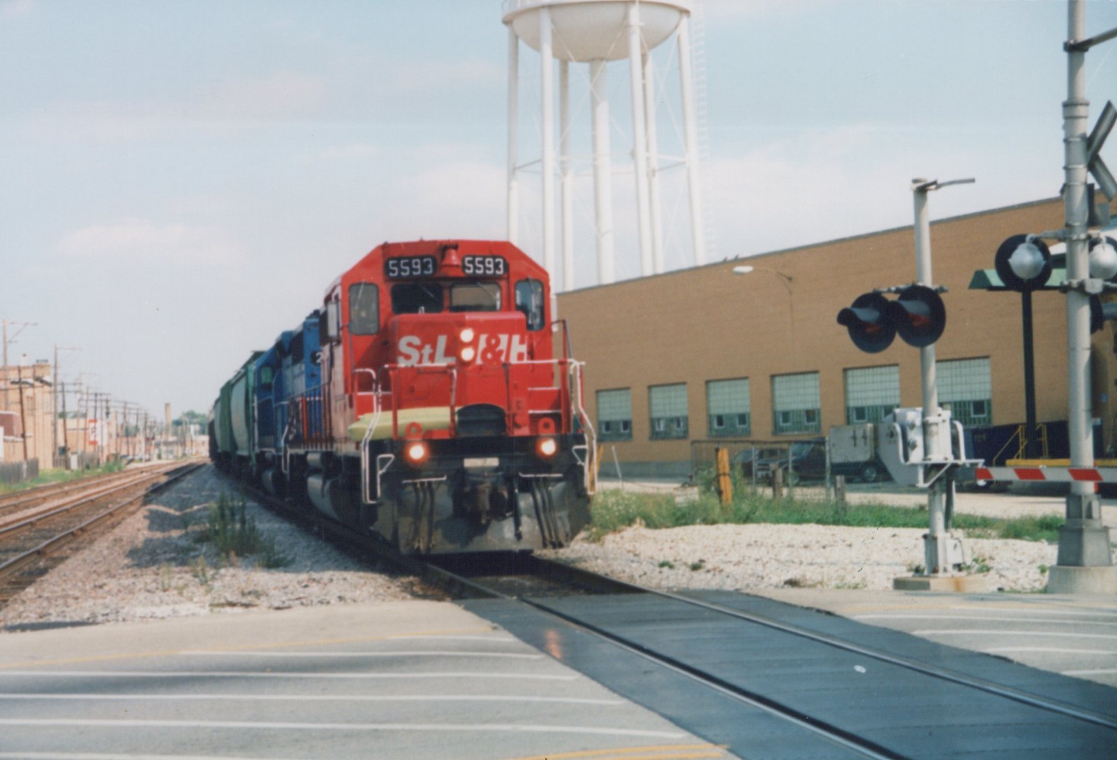 A red and yellow diesel locomotive with the number 5593 and StL&H lettering is moving on tracks across a street, with a brick building and a white water tower in the background.