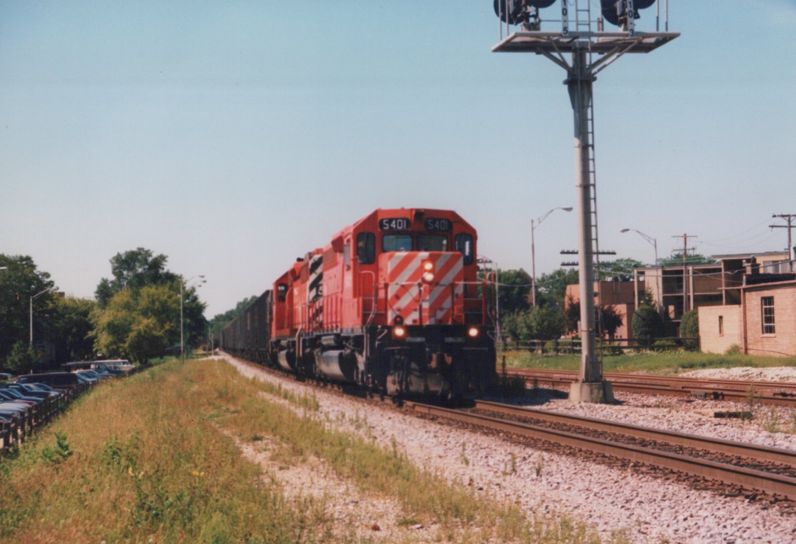A red Canadian Pacific locomotive numbered 5401 is pulling a long freight train on a sunny day, viewed from the side of the tracks with houses and trees in the background.