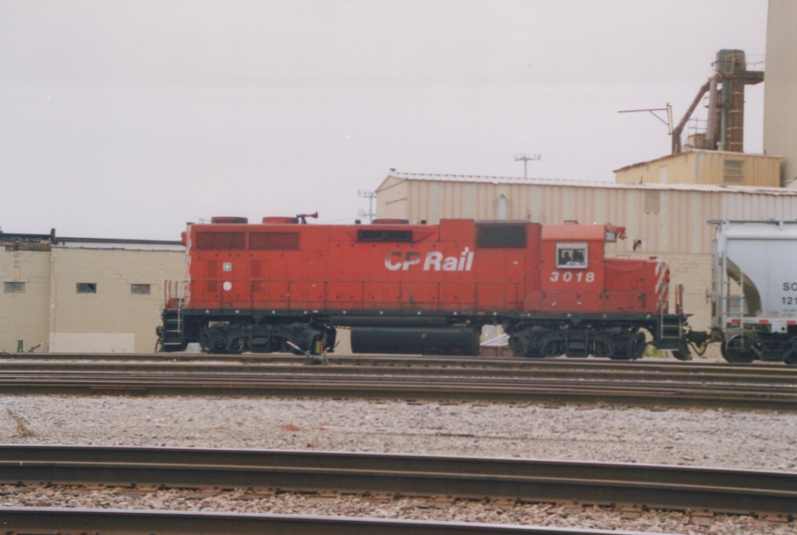 A red CP Rail SD40-2 diesel locomotive, number 3018, on a railroad track next to a white freight car, with industrial buildings in the background under an overcast sky.