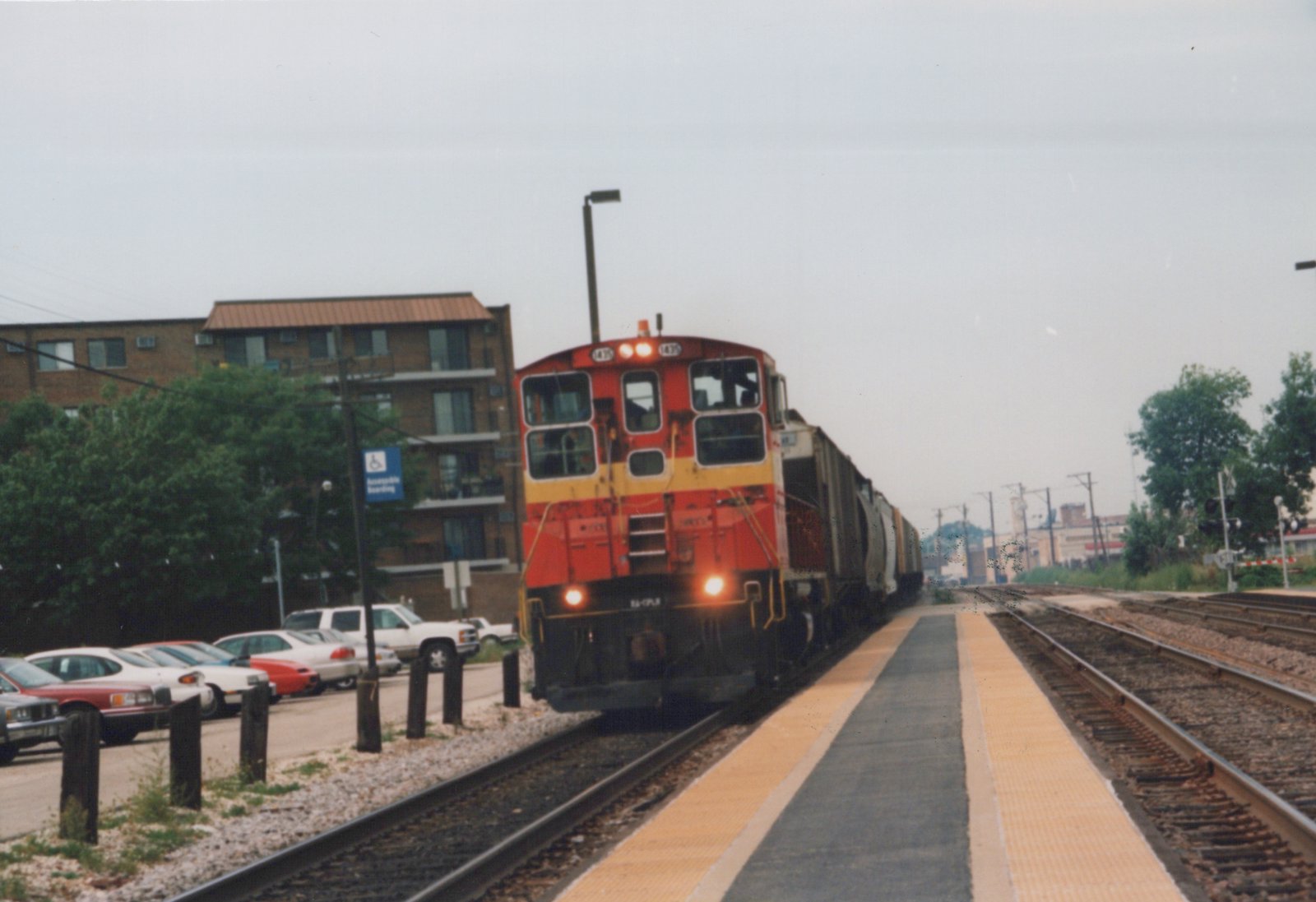 A red and yellow diesel locomotive with the number 475 on its front is moving along tracks next to a platform. Cars are parked on the left, and a multi-story building is in the background under a cloudy sky.