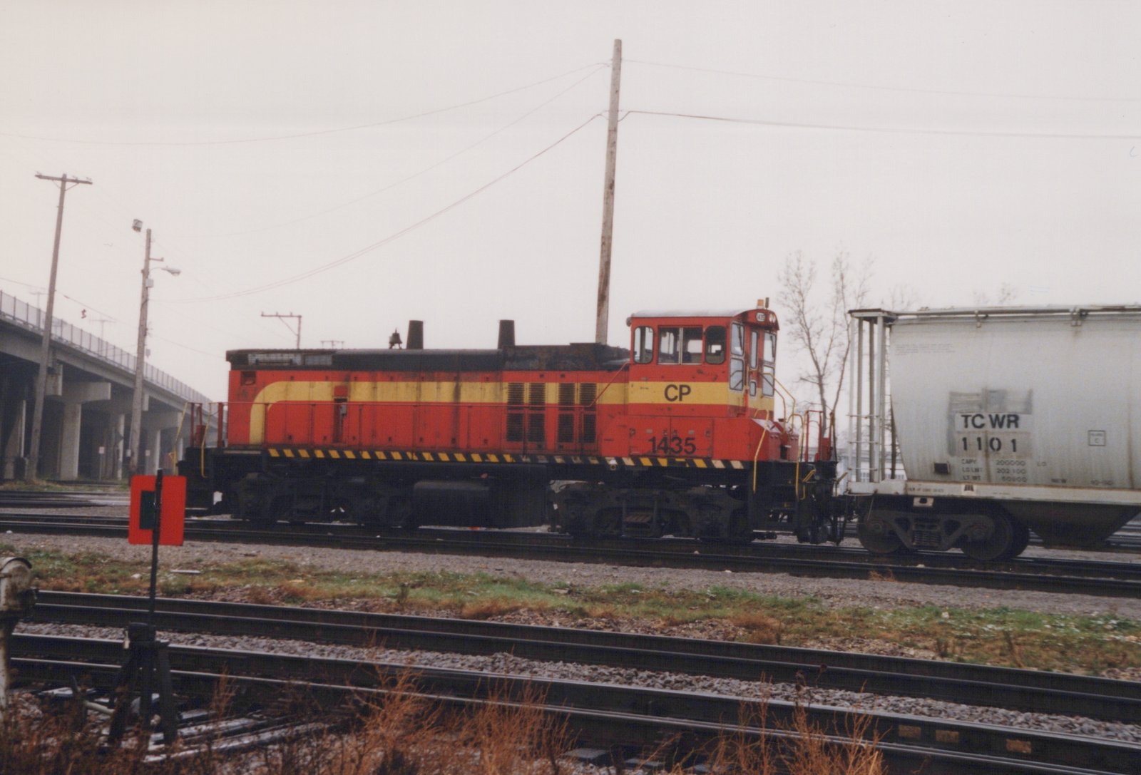 Side view of a red and yellow Canadian Pacific locomotive number 1435 coupled to a white covered hopper car, number TC WR 1101, on multiple tracks in a rail yard with a bridge and bare trees in the background.