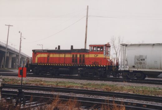 Side view of a red and yellow Canadian Pacific locomotive number 1435 coupled to a white covered hopper car, number TC WR 1101, on multiple tracks in a rail yard with a bridge and bare trees in the background.
