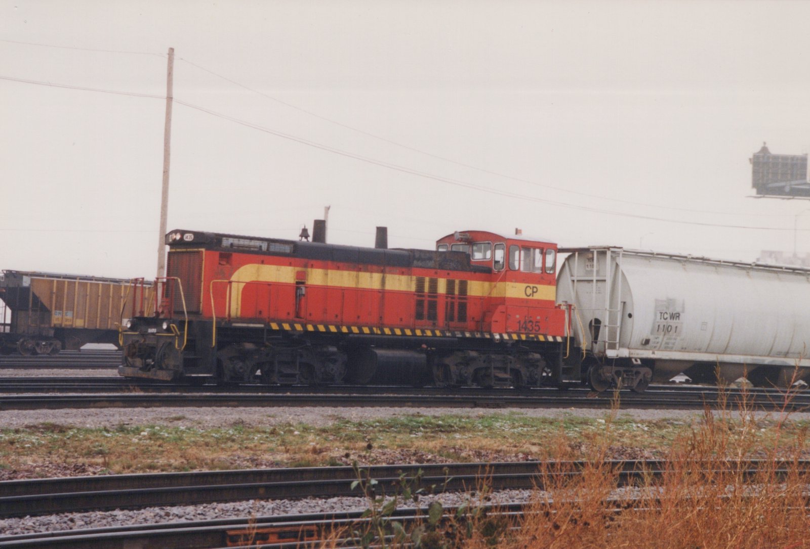 A red and yellow Canadian Pacific GP9 diesel locomotive, number 1435, is parked in a rail yard with a white TCWR tank car and a brown ON boxcar nearby.