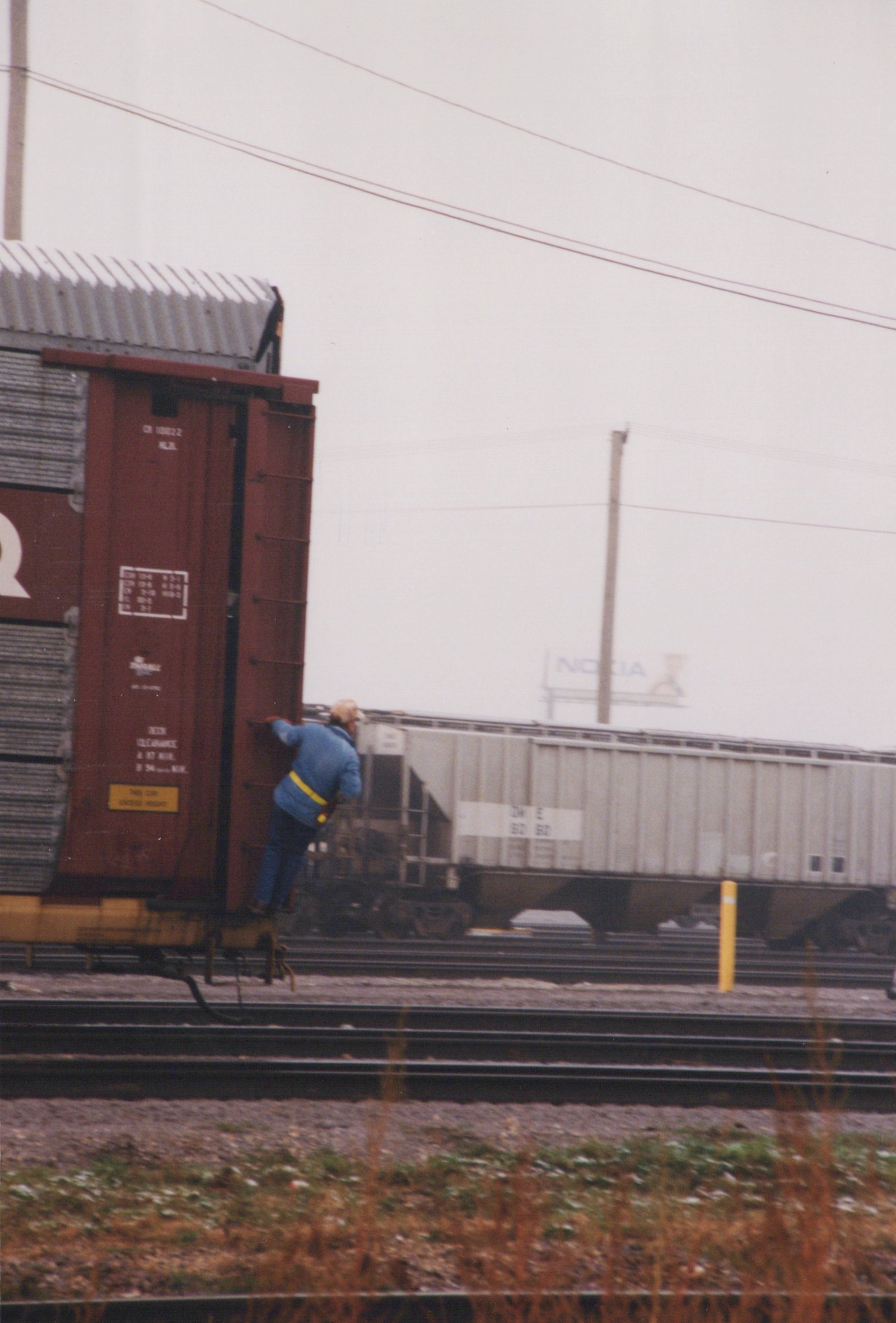 A worker in a blue jacket climbs onto a red Chicago and North Western boxcar, with a gray covered hopper and a Nokia billboard in the background.