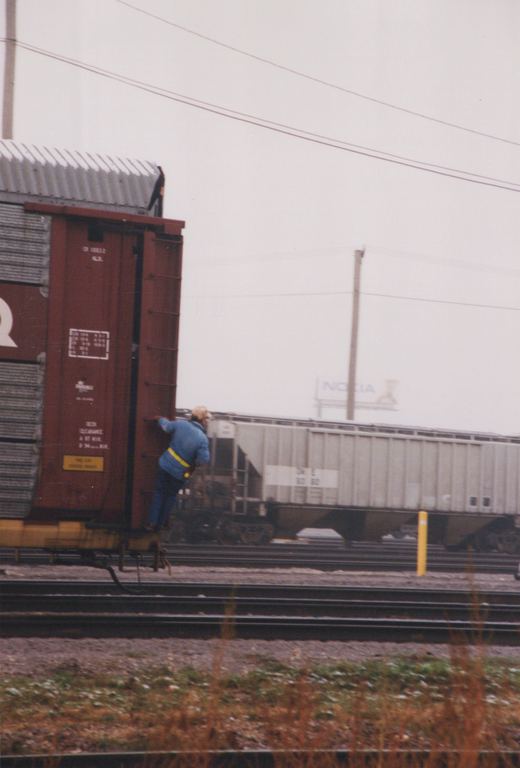A worker in a blue jacket climbs onto a red Chicago and North Western boxcar, with a gray covered hopper and a Nokia billboard in the background.