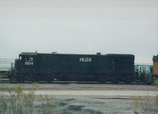 A black HLGX locomotive with the number 6814 on its side is shown in profile, sitting on railroad tracks in a yard with other freight cars and overgrown weeds in the foreground.