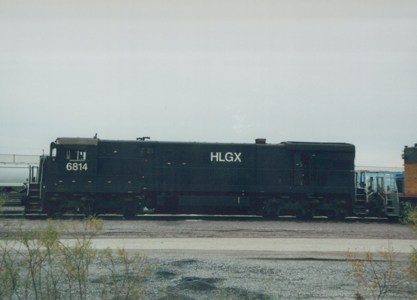 A black HLGX locomotive with the number 6814 on its side is shown in profile, sitting on railroad tracks in a yard with other freight cars and overgrown weeds in the foreground.