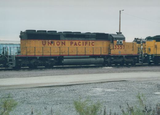 A side view of three yellow and gray Union Pacific locomotives, numbers 3357, 2966, and 4307, in a gravel yard with a blue freight car visible to the left.