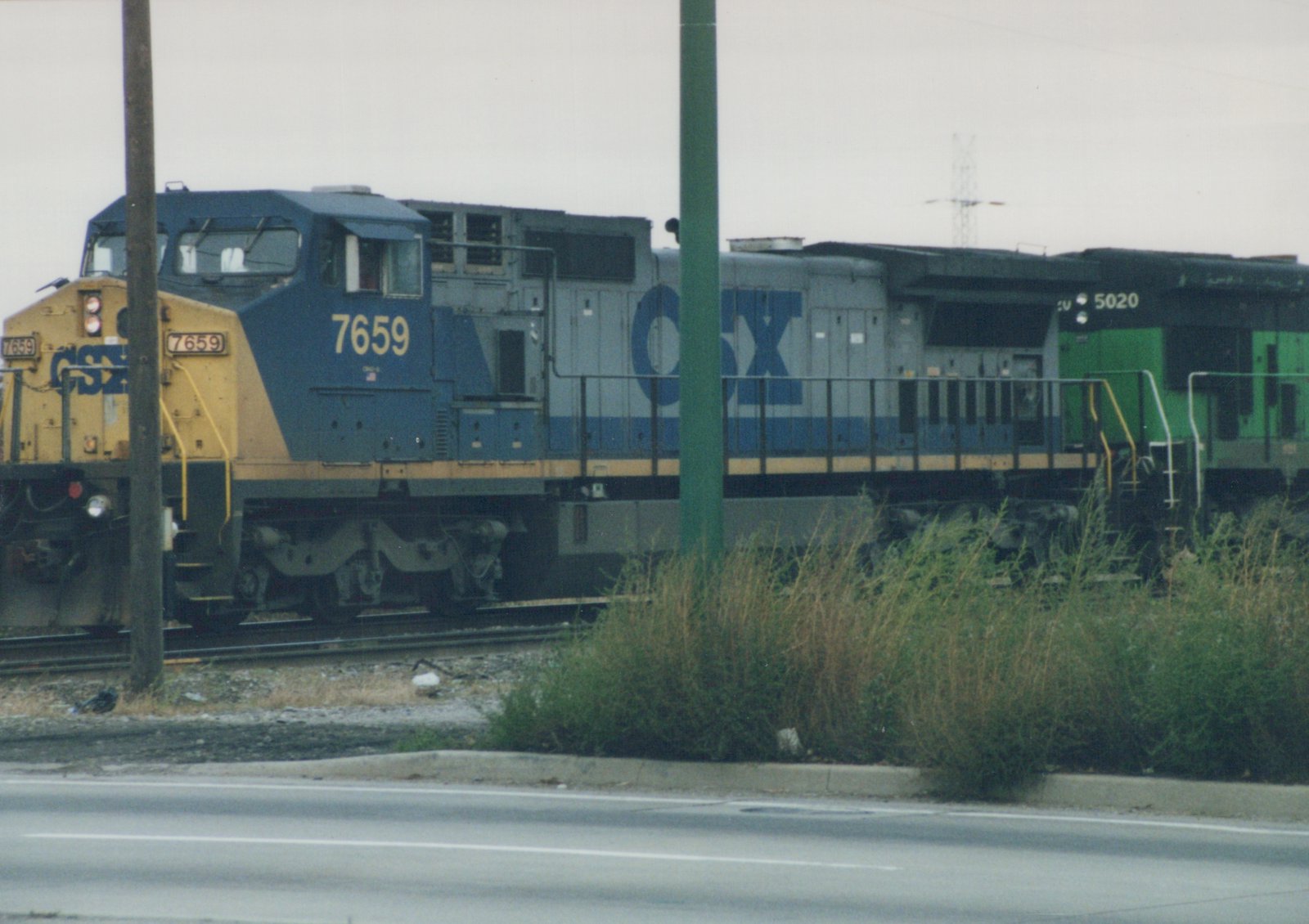 A side view of a blue and yellow CSX diesel locomotive numbered 7659, coupled with another green and black CSX locomotive numbered 5020, on a railroad track next to a road with tall grass in the foreground.