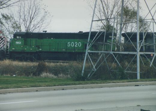 A side view of a green RMGX diesel locomotive numbered 5020 on a railroad track beside a paved road, with metal transmission structures nearby and leafless trees in the background.