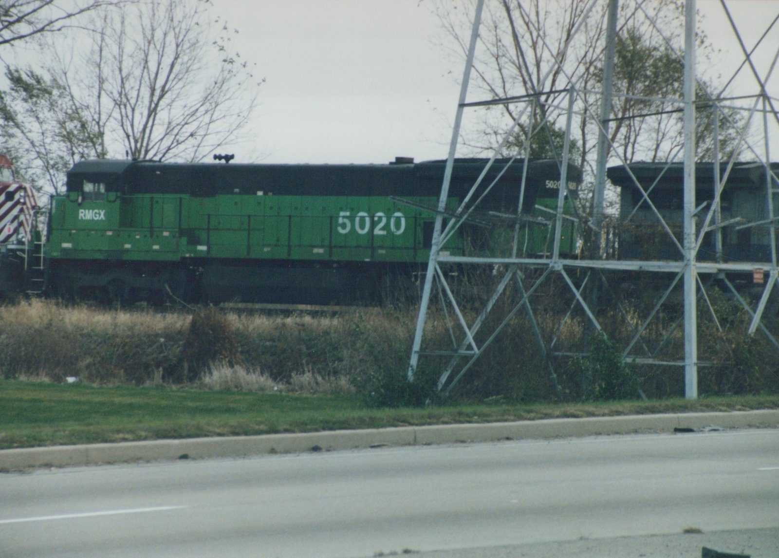 A side view of a green RMGX diesel locomotive numbered 5020 on a railroad track beside a paved road, with metal transmission structures nearby and leafless trees in the background.