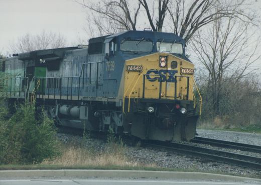 A blue and yellow CSX diesel locomotive, number 7659, is shown on railroad tracks next to a road, with bare trees in the background on an overcast day.