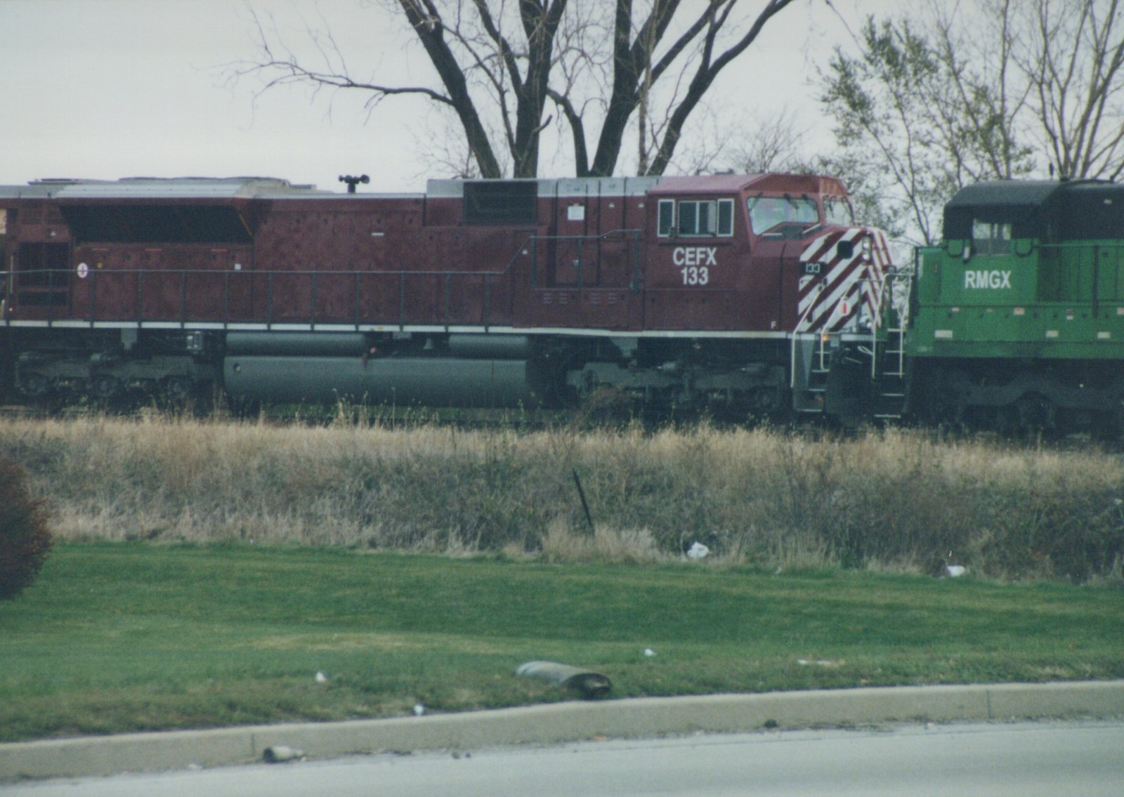 A red CEFX locomotive numbered 133 is shown on a track beside a green RMGX locomotive, with an overcast sky and leafless trees in the background.