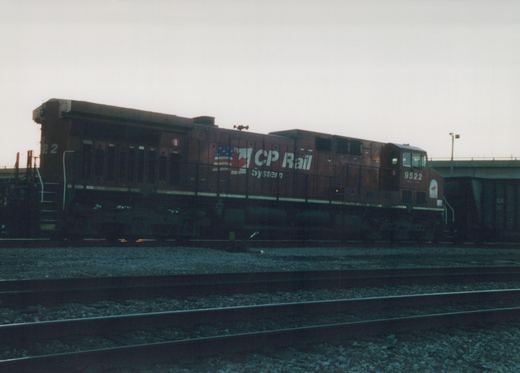 Side view of a maroon CP Rail System diesel locomotive, number 9522, on a track with multiple rails and gravel ballast, with a bridge in the background under an overcast sky.