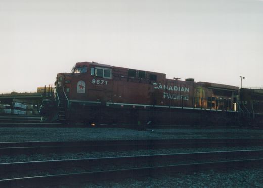 A red Canadian Pacific diesel locomotive, number 9671, sits on multiple tracks in a rail yard at dusk, with a second locomotive, number 822, visible behind it.