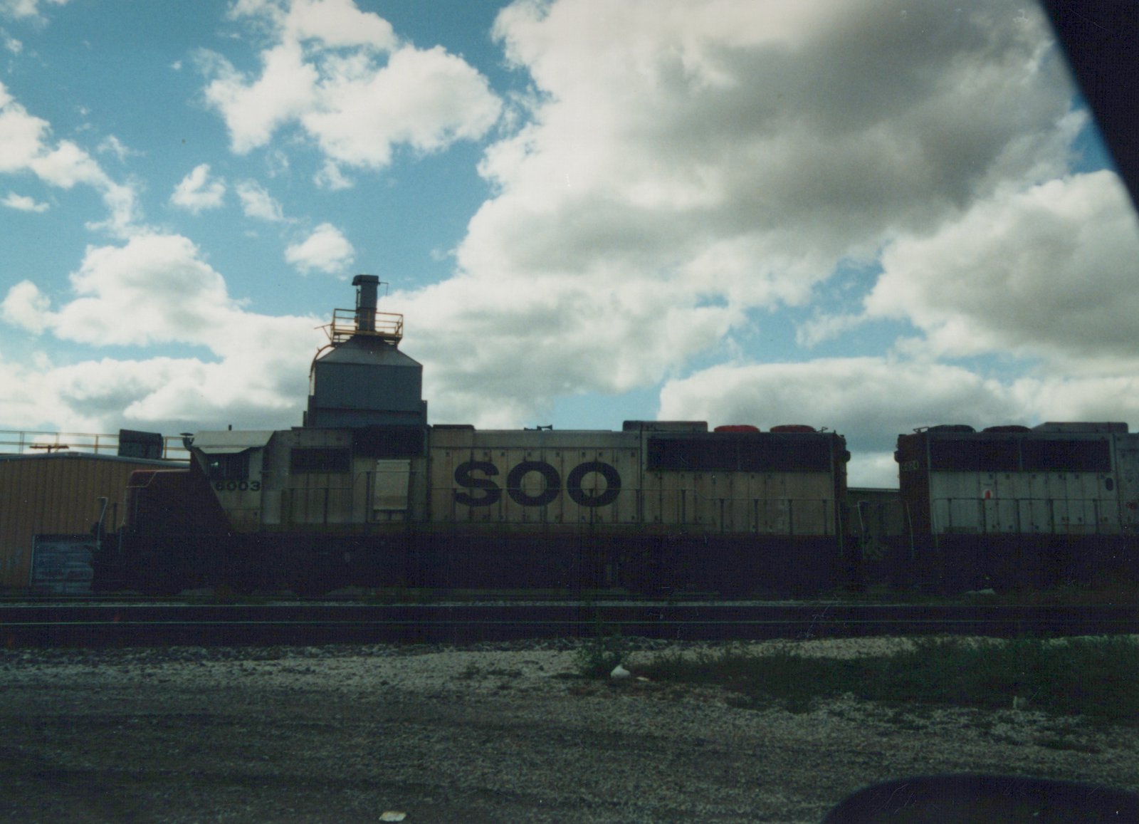 Two gray and black Soo Line diesel locomotives, numbered 6003 and 6024, are parked on parallel tracks in a gravel yard with a large industrial building to the left and a cloudy sky above.