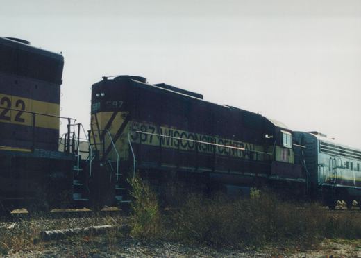 A dark blue and yellow Wisconsin Central locomotive with the number 587 on its side is parked on tracks in a rail yard with other railcars nearby.