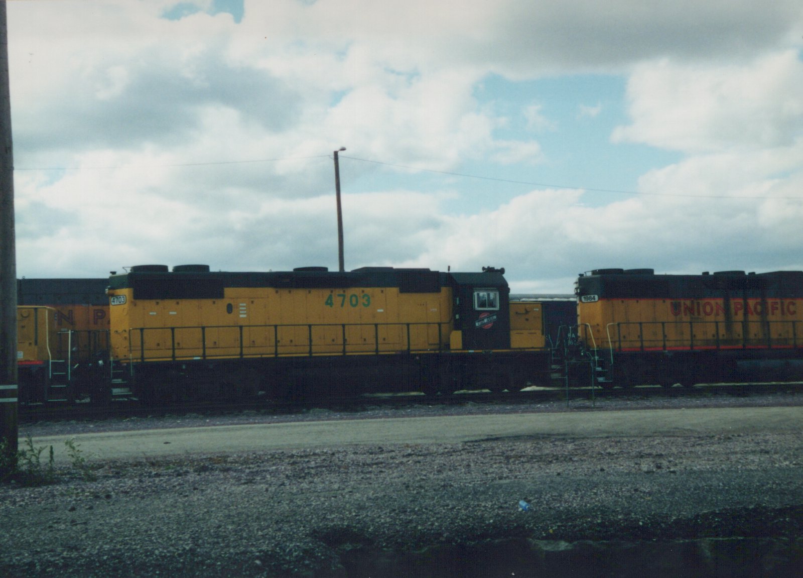 Two yellow diesel locomotives, one marked Chicago and North Western 4703 and the other Union Pacific 1984, are parked on parallel tracks in a gravel yard under cloudy skies.