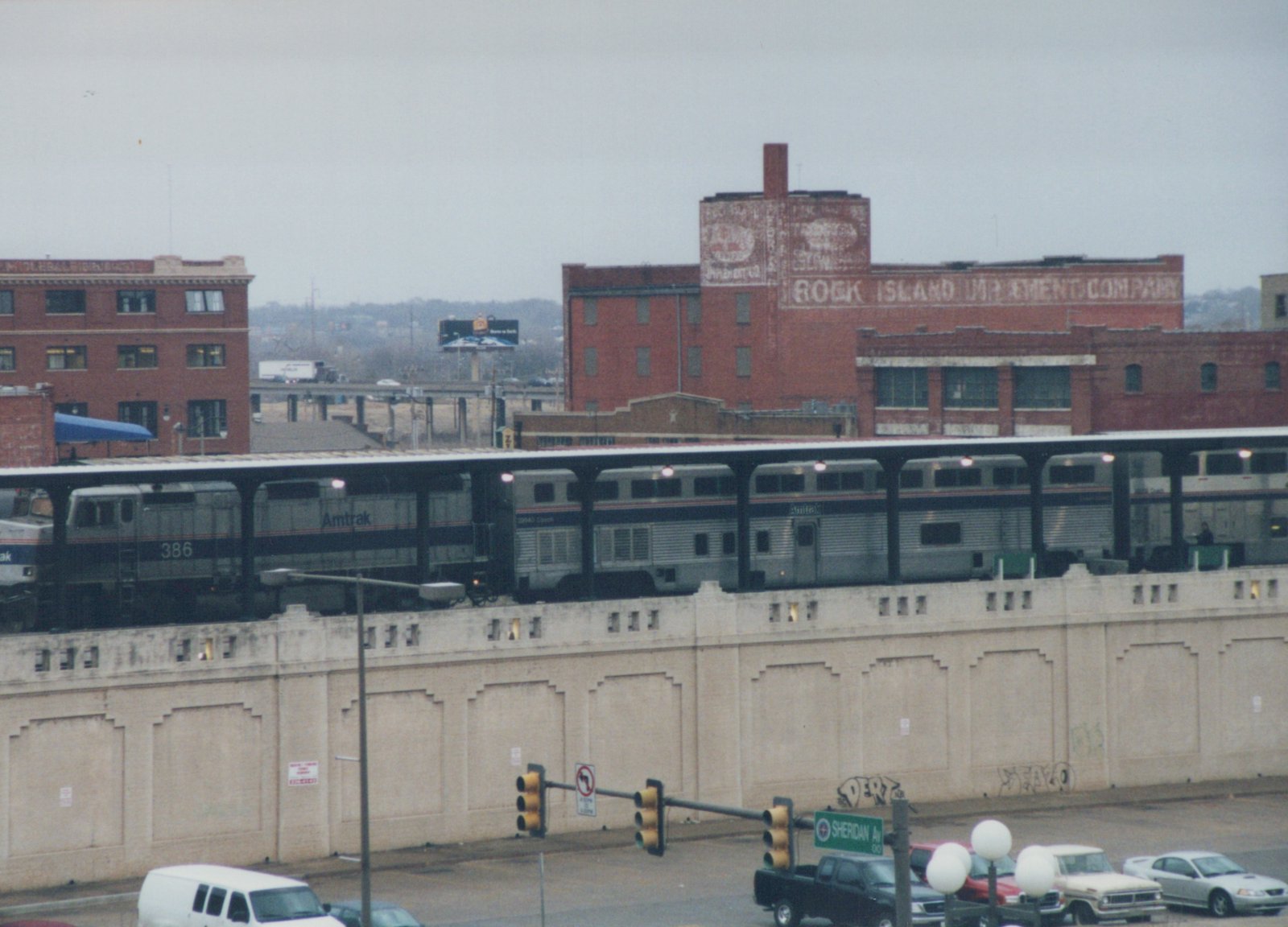 An Amtrak train with locomotive number 386 on an elevated platform, passing by brick buildings and street traffic below on a winter day.