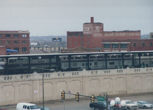 An Amtrak train with locomotive number 386 on an elevated platform, passing by brick buildings and street traffic below on a winter day.