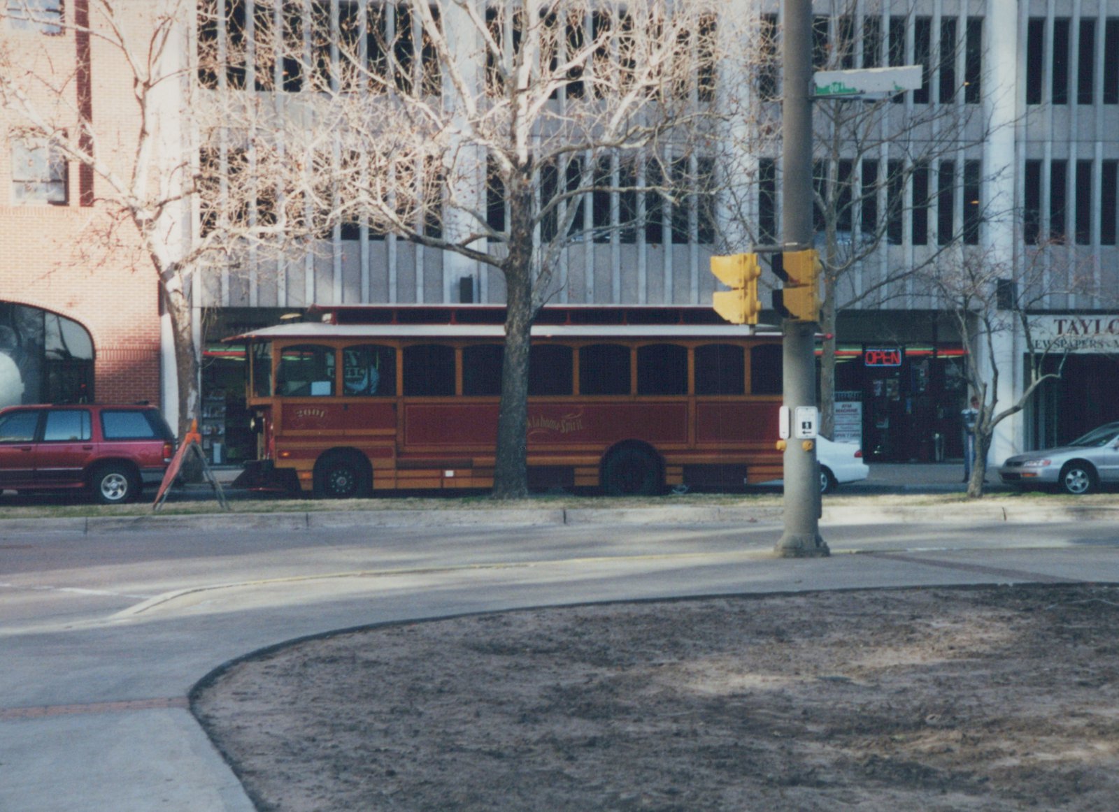A maroon and gold motor bus with the number 2007 and 'Oklahoma Spirit' lettering is parked on a city street with bare trees and other cars nearby.