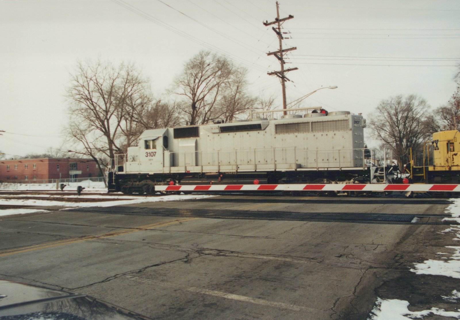 A white diesel locomotive with the number 3107 and CEFX lettering is stopped at a grade crossing with red and white barriers. Snow is on the ground, and bare trees are in the background.
