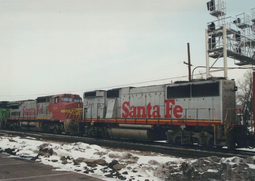 Two Santa Fe diesel locomotives, one red and one grey, are parked on tracks beside a snow-covered road, with a large signal tower in the background.
