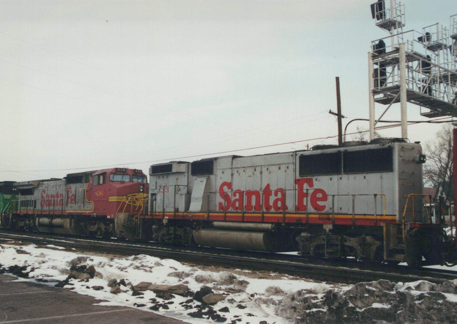 Two Santa Fe diesel locomotives, one red and one grey, are parked on tracks beside a snow-covered road, with a large signal tower in the background.