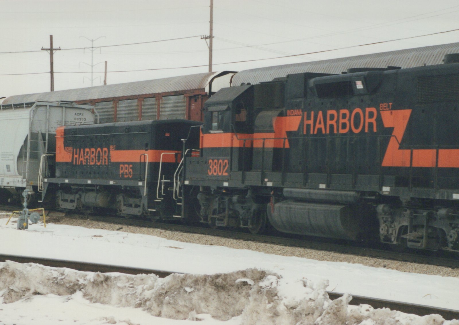 A black and orange Indiana Harbor Belt diesel locomotive numbered 3802 sits on tracks in a snow-covered yard, with a gray freight car behind it.