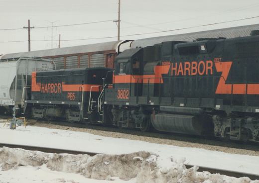 A black and orange Indiana Harbor Belt diesel locomotive numbered 3802 sits on tracks in a snow-covered yard, with a gray freight car behind it.