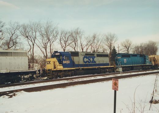 A blue and yellow CSX freight train with two locomotives, numbers 8450 and 6345, travels on tracks beside snow-covered ground with bare trees in the background.