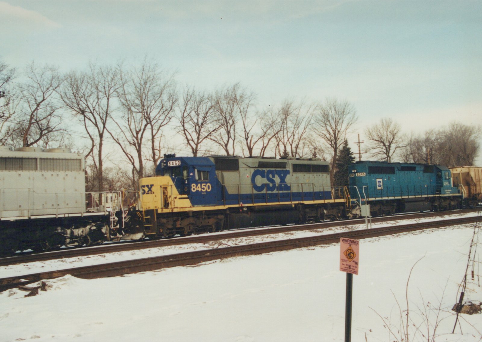 A blue and yellow CSX freight train with two locomotives, numbers 8450 and 6345, travels on tracks beside snow-covered ground with bare trees in the background.