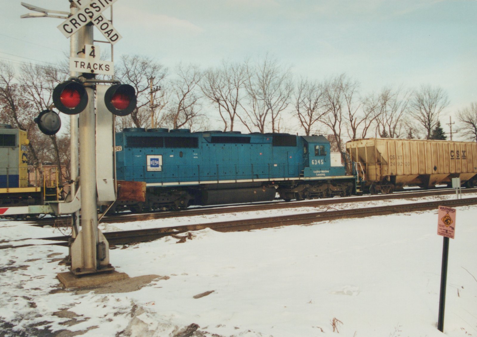 A blue Electro-Motive Leasing diesel locomotive numbered 6345 pulls a beige CSX hopper car past a railroad crossing with warning lights and signs, set against a snowy landscape with bare trees.