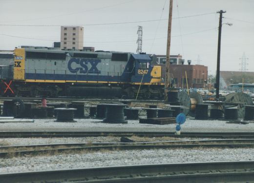 A side view of a blue, gray, and yellow CSX diesel locomotive numbered 8247, sitting on tracks in a yard with cable spools and buildings behind it on an overcast winter day.
