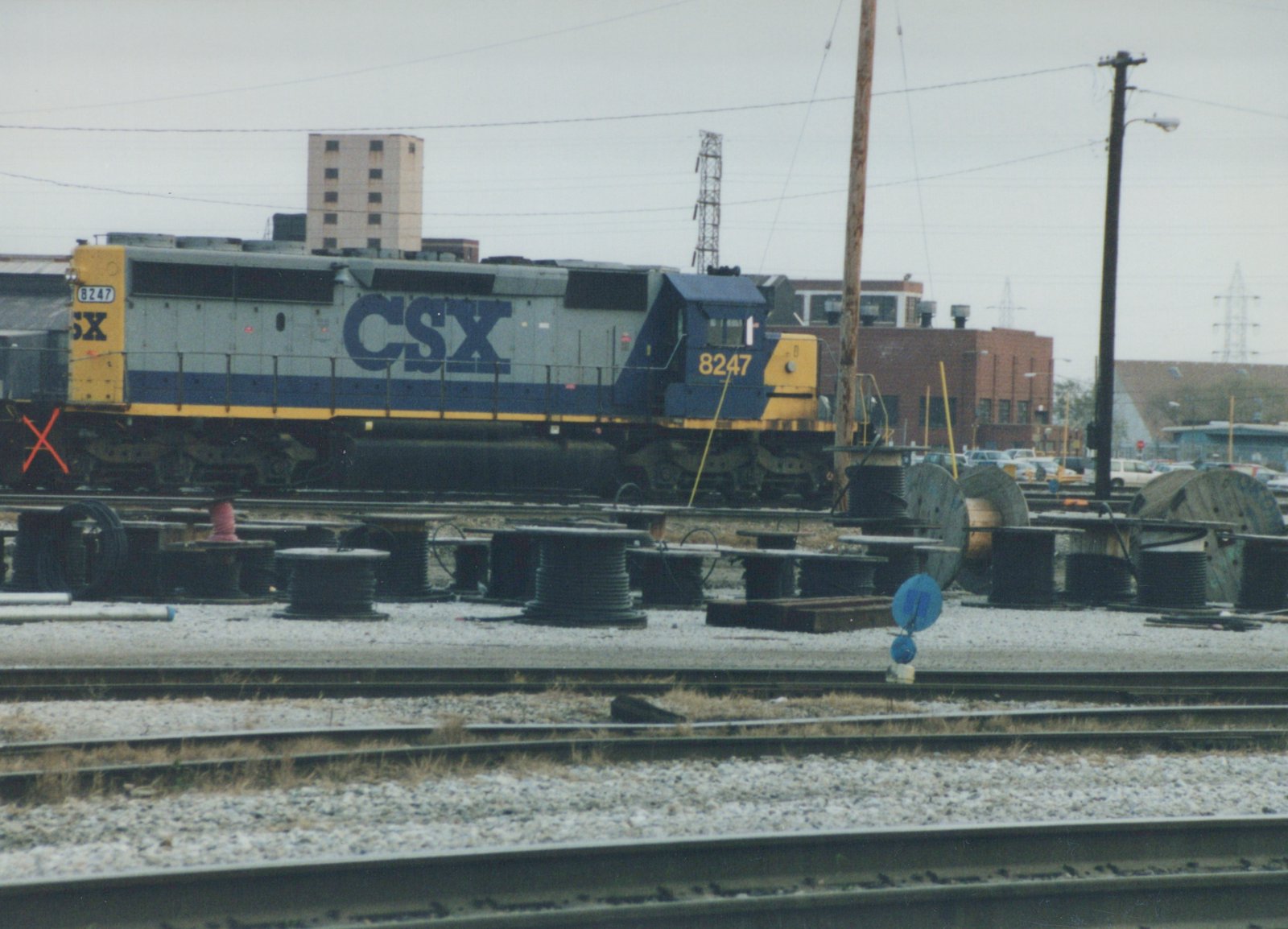 A side view of a blue, gray, and yellow CSX diesel locomotive numbered 8247, sitting on tracks in a yard with cable spools and buildings behind it on an overcast winter day.