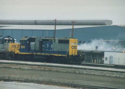 A blue and yellow CSX diesel locomotive numbered 2614 is on railroad tracks in a snow-dusted yard, with another CSX locomotive numbered 1517 behind it, under an overcast sky.